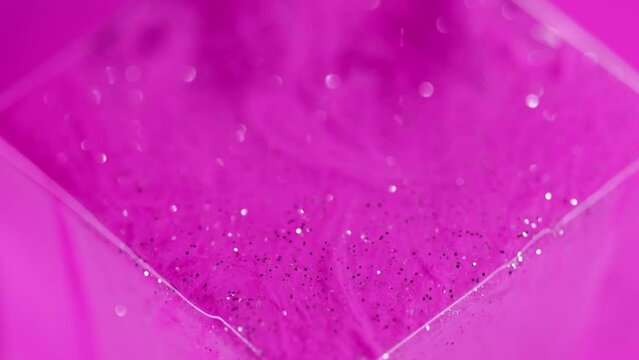 Glitter ink. Shimmering spill. Bokeh light. Defocused bright pink color shiny silver sequin particles texture gel fluid paint flow on glass cube geometric art abstract background.