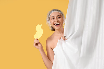 Young woman in shower cap with paper duck behind bathroom curtain on yellow background