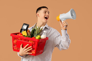 Young man with full shopping basket and payment terminal shouting into megaphone on beige background