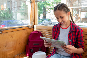 A brunette girl of 12-14 years old, a traveler with a backpack, riding in an old tram, looking at a map and sights. Vacation, travel, trip and summer is the concept of summer travel.