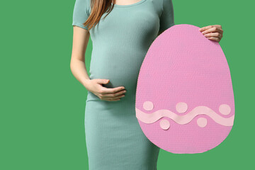 Young pregnant woman with paper Easter egg on green background