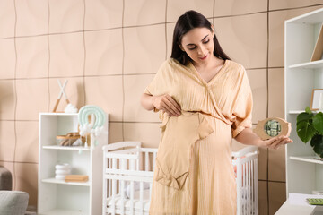 Young pregnant woman with baby bodysuit and piggy bank at home. Maternity Benefit concept