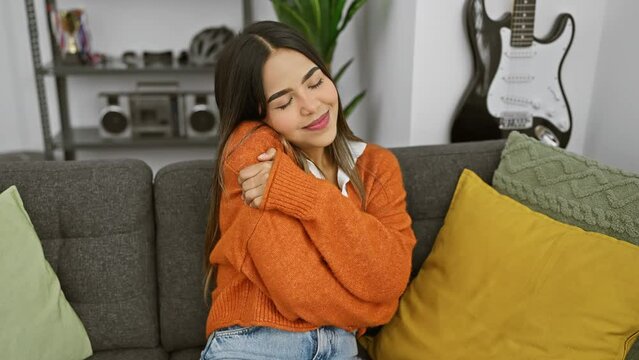 Young beautiful latin woman wearing sweater sitting on sofa hugging oneself happy and positive, smiling confident. self love and self care at home