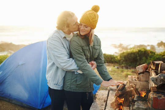 Happy couple, kiss and bonding together by campfire, outdoor and romantic getaway with camping in forest. Man, woman and love on vacation on weekend break, countryside and travel in woods for sunset