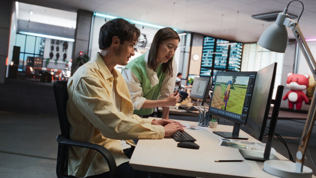 Caucasian Male Game Designer Using Desktop Computer With 3D modelling Software And Talking to Asian Female Art Director in Game Development Studio Office. Diverse Colleagues Discussing New RPG Game