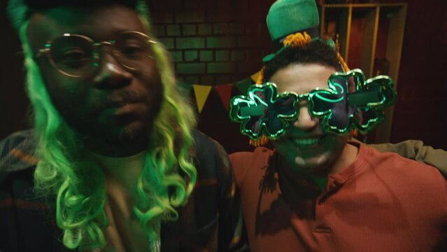UGC POV handheld footage of group of multiethnic friends wearing green wigs and funny glasses and celebrating Saint Patricks Day in bar while dancing near table with glasses of green beer