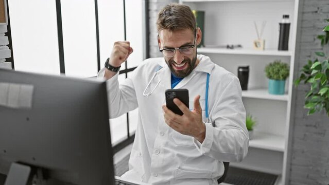 Ecstatic young hispanic man, a handsome bearded doctor, celebrating a win in the clinic while texting on his smartphone