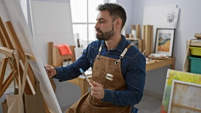 Young handsome hispanic man artist concentrates, draped in apron, beard highlighted, interiors of studio buzz with focus, drawing class at art academy, learning the brush strokes on canvas.