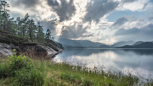 Sun rays pierce through the stormy clouds whirling over the lake and a forest in a timelapse video.