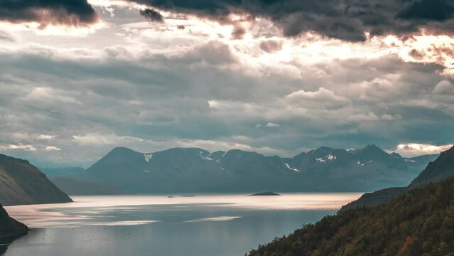 Stormy clouds backlit by the setting sun whirl above the calm fjord and mountains in the timelapse video.