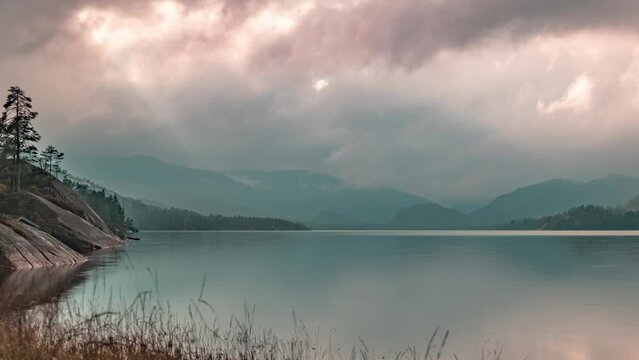 Piercing sun rays shine through the stormy clouds whirling over the lake with rocky banks and mountains in a timelapse video.