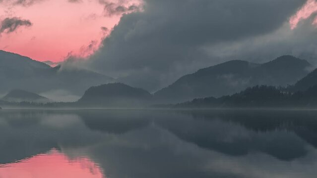 Heavy clouds in the pink sunset sky reflected in the mirrorlike water. Dark mountains wrapped in fog tower in the background. Timelapse video.