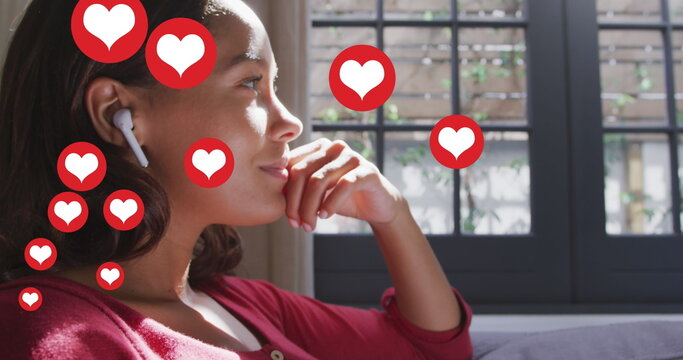 Image of digital heart icons over woman listening to music with earphones writing at home