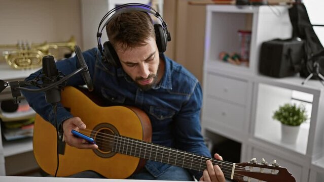 Handsome young hispanic man sitting in a music studio, passionately playing a chord on a classical guitar while looking at his smartphone screen. a melodious artist lost in musical world.