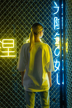 A beautiful blonde girl in a white oversized T-shirt and blue jeans posing against the background of neon hieroglyphs