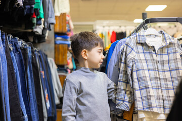 happy preschooler boy kid in clothes store is shopping, choosing. happy smile child with shirt or sport suit on hangers in hand. many, lot of different colorful clothes in shop for kids.