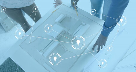 Image of lightbulb icons over diverse colleagues with blueprints in office