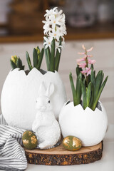 Happy Easter. A spring hyacinth flower in an egg-shaped vase, Easter bunnies and eggs with a golden pattern on the table. There is a white kitchen in the background. Easter decor in the house.