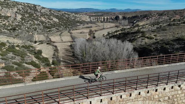 Aerial lateral dolly scene of a cyclist crossing a  old bridge of an abandoned mining railway, Via Verde Ojos Negros, Teruel, Spain.4K 60FPS