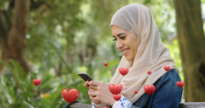Muslim woman in hijab texts with hearts flying around her on a bench in 4k. - Powered by Adobe