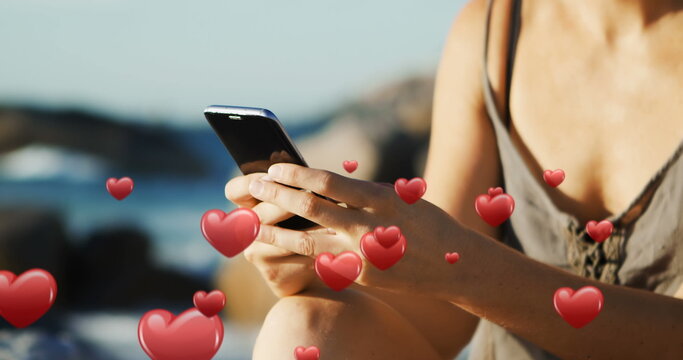 Woman texts by the beach with digital hearts around in 4K. - Powered by Adobe