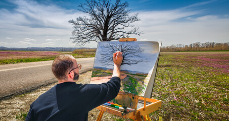 Male artist paints large lonely oak tree growing in filed by rural road early in spring; fields and...