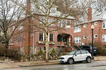 Traditional two-story red brick house surrounded by bare trees, with a parked car on the street in...