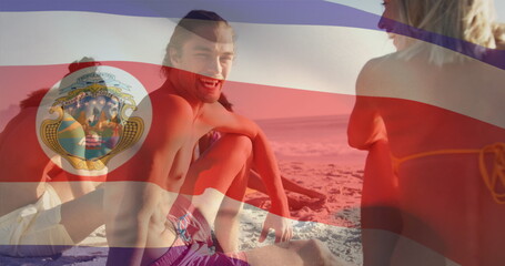 Image of flag of costa rica waving over diverse friends sitting and enjoying at beach