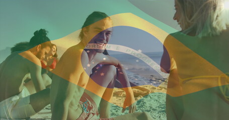 Image of flag of brazil waving over diverse friends discussing while sitting on beach