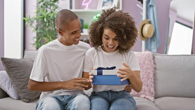 Beautiful couple enjoy fun times at home, the man covering the woman's eyes on the sofa for a lovely birthday surprise gift, smiling with confident love.