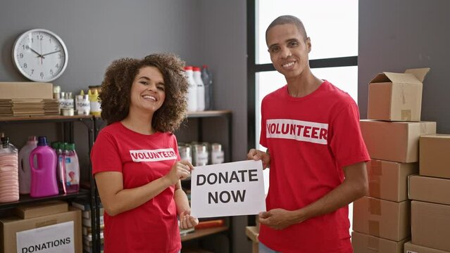 Man and woman volunteers, smiling together, holding 'donate now' banner at charity center, showing unity in altruism while working indoors