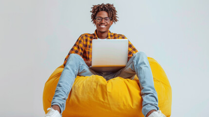 A joyful person sits cross-legged on a yellow beanbag, working on a laptop with a smile. - obrazy, fototapety, plakaty