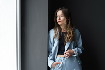 Contemplative young woman in a chic blue suit standing beside a window.