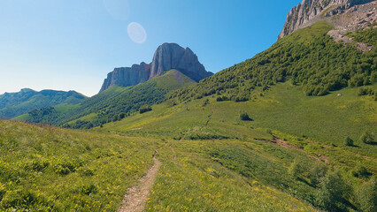 Natural mountain scenery. The trail passes through green grass. A majestic cliff rises ahead.