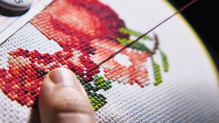 A close-up of a hand delicately performing cross-stitch embroidery, creating a colorful and intricate design on fabric held taut by a hoop frame.