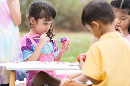Happy group of cute little diverse children painting Easter eggs together. Kid girl holding painting brush, painting watercolor on egg while playing outdoors at park
