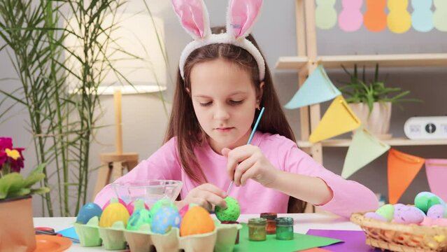 Concentrated little child wearing bunny ears headband painting easter eggs at home drawing beautiful red spots on green egg preparing for spring traditional holiday celebration