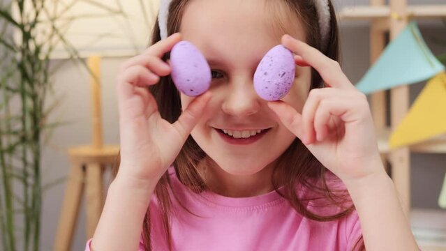 Extremely happy brown haired little girl wearing bunny ears headband playing with purple decorating eggs covering eyes and smiling with happiness being in good festive mood