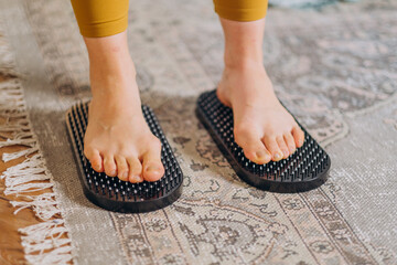 A pair of feet stand on a black acupressure mat placed on a traditional rug, highlighting an at-home wellness practice. The mat's spikes are designed for foot stimulation and relaxation.