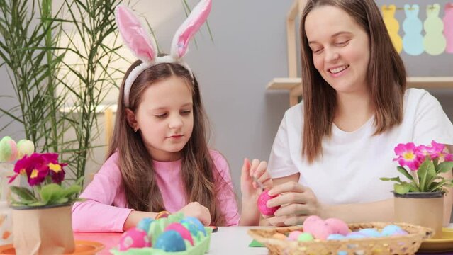 Easter preparation activity. Brown haired little girl with mother painting Easter eggs in festive home interior young woman enjoying happy activities during holiday celebration