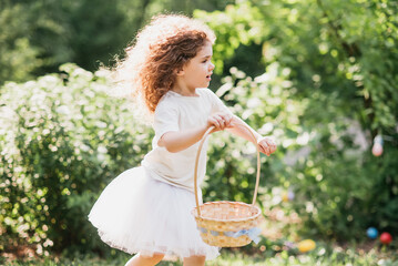 Easter egg hunt. Girl child Wearing Bunny Ears Running To Pick Up Egg In Garden. Easter tradition. Baby with basket full of colorful eggs. - 757832342