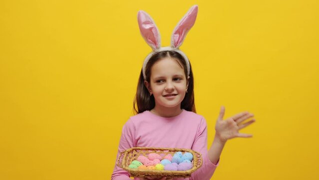 Cute little girl wearing pink bunny ears headband holding painted eggs in basket standing isolated over yellow background holding hand near mouth making announcement inviting to holiday party