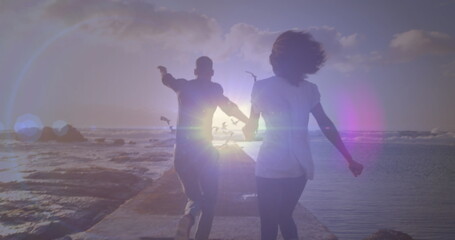 Bright spot of light against rear view of african american couple running on the rocks near the sea - Powered by Adobe