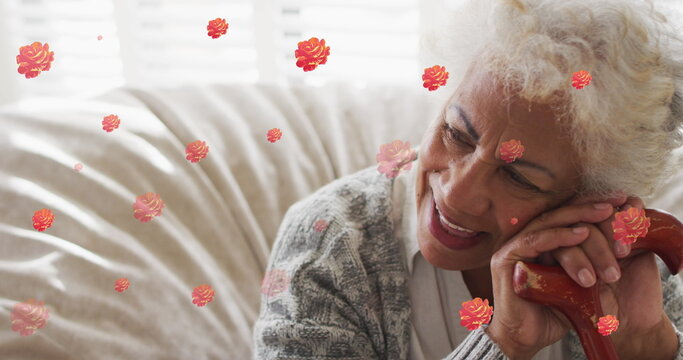 Image of red roses over smiling biracial senior woman sitting leaning on walking stick, at home