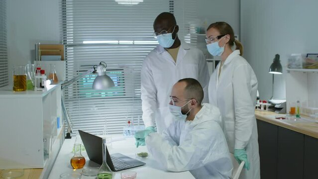 Medium shot of multiracial team of scientists in lab coats and face masks looking at laptop screen and having discussion while working on research together in modern laboratory