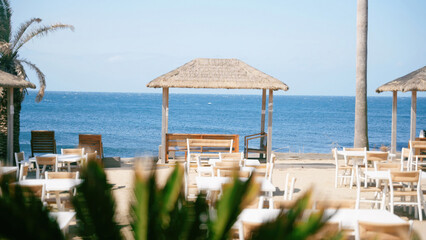 A beach restaurant or bar with tables, chairs, and thatched umbrellas, overlooking a calm blue sea.