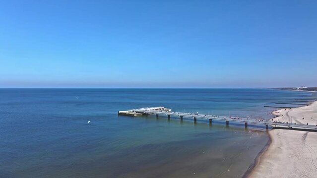 Aerial view of Kołobrzeg pier, beach, tourists, blue sky, and seabirds in February drone video.