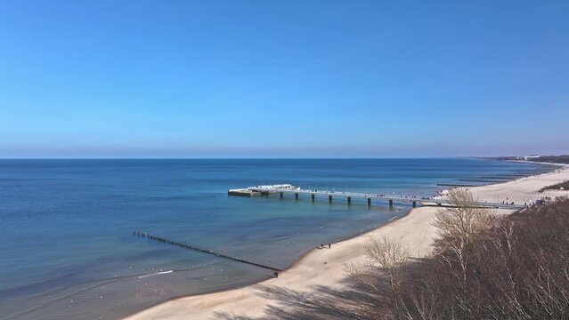 Sunny February day in Kołobrzeg: drone records pier, beach, tourists, blue sky, seabirds.