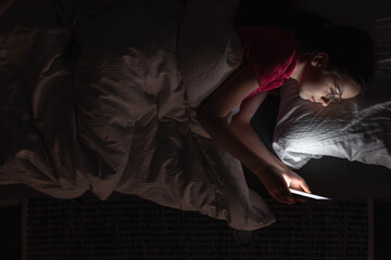 A teenage girl with a smartphone lies in bed at night, top view.
