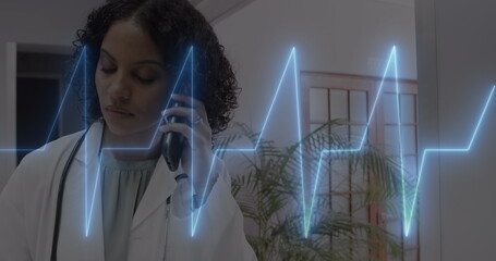 Image of cardiograph over happy biracial female doctor talking on smartphone